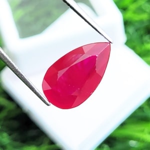 May include: A pear-shaped, faceted gemstone with a vibrant, deep red color. The stone is held by a pair of silver-toned metal tweezers, showcasing its clear, polished surface and precise cut against a bright, reflective background.