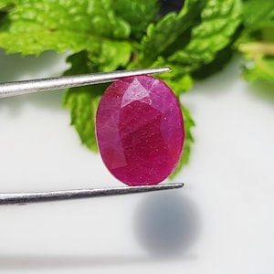 Op de afbeelding: Een ovaalvormige, gefacetteerde robijn met een diepe magenta-rode kleur. De edelsteen wordt stevig vastgehouden door een zilverkleurige metalen pincet, waardoor het gepolijste oppervlak en de geometrische slijpvorm tegen een lichte achtergrond zichtbaar zijn.