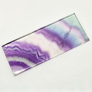 Puede incluir: Una losa rectangular de cristal de fluorita con una superficie pulida. La piedra presenta un patrón en capas de tonos morados, blancos y azules. Los bordes son claros y la forma general es un rectángulo largo. El cristal está sobre un fondo blanco.