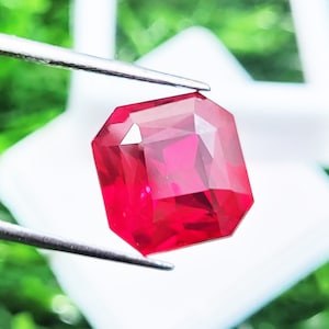 May include: A vibrant, deep red gemstone with a faceted, rectangular cut held by silver-toned metal tweezers. The stone displays sharp geometric edges and a polished surface that reflects light, showcasing a rich, saturated crimson color throughout.