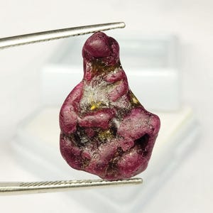 May include: A raw, unpolished ruby gemstone with a deep magenta and dark red colour palette. The stone features an irregular, organic shape with visible surface textures, natural crevices, and subtle golden-brown inclusions throughout its structure.