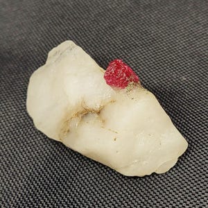 May include: A raw, translucent white mineral specimen featuring a single, vibrant red crystal formation embedded on its surface. The white stone has an irregular, jagged shape with subtle golden-coloured mineral inclusions running through its centre.