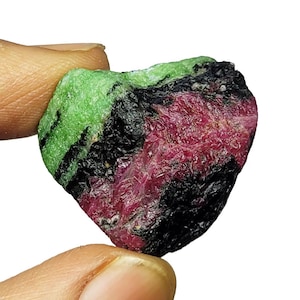 May include: A raw, unpolished mineral specimen featuring a vibrant combination of colors. The stone displays a bright green section, a deep black area with a rough texture, and a prominent patch of crystalline, deep magenta-red mineral matter.