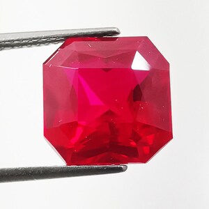 May include: A vibrant, deep red gemstone with a square, faceted cut held by silver-toned metal tweezers. The stone exhibits high clarity and internal light reflection, showcasing a sharp, geometric shape with beveled edges and a polished surface.