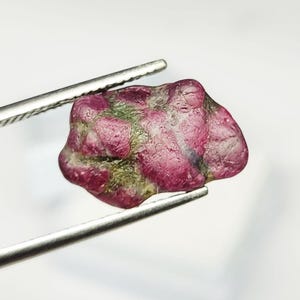 May include: A raw, unpolished gemstone featuring a mix of deep magenta and dark olive green mineral deposits. The stone has an irregular, rounded shape with a textured surface and is held by a pair of silver-toned metal tweezers.