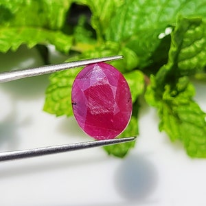 May include: A vibrant, oval-cut ruby gemstone with a deep pinkish-red hue and a faceted surface. The gemstone is held by a pair of silver-toned metal tweezers, showcasing its translucent quality and internal structure against a bright backdrop.