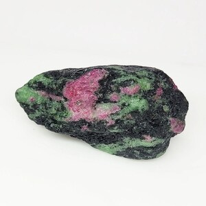 Op de afbeelding: Een ruw, ongepolijst mineraal exemplaar met een mix van levendige magenta-roze, diep bosgroene en donkerzwarte kristallijne texturen. De steen heeft een onregelmatige, langwerpige vorm met een ruw, natuurlijk oppervlak met kleurzones.
