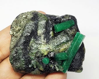 Könnte beinhalten: Ein rohes Mineralexemplar mit leuchtend grünen Smaragdkristallen, die in eine dunkle, texturierte Matrix eingebettet sind. Der Stein enthält metallische Pyriteinschlüsse auf einer Seite und mehrere ausgeprägte, längliche, hexagonale Smaragdprismen.