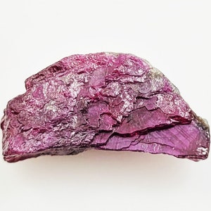 May include: A raw, deep magenta-coloured gemstone with a rough, layered, and crystalline texture. The stone features a semi-circular shape with jagged, uneven edges and a reflective, multifaceted surface that catches light across its dark pink body.