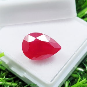May include: A vibrant, pear-shaped, red gemstone with multiple facets. The gem is presented in a white box, set against a green backdrop. The cut of the gem creates many reflective surfaces, enhancing its sparkle. This gemstone is likely for jewellery.