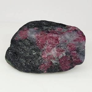 Puede incluir: Una gema de rubí en bruto, sin cortar, con un exterior oscuro y texturizado. La piedra presenta parches de cristales rojos profundos y vibrantes sobre un fondo gris y negro. La forma irregular resalta la belleza natural y sin pulir del mineral.