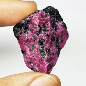 Può includere: Un esemplare minerale grezzo e non lucidato caratterizzato da un vivace colore di base rosa magenta con inclusioni nere irregolari e sparse in tutta la struttura cristallina. La pietra presenta una superficie ruvida e testurizzata con bordi frastagliati.