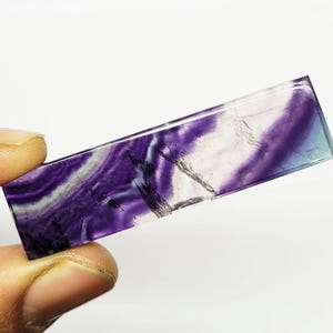 Puede incluir: Un cristal rectangular de fluorita con una superficie pulida. La piedra presenta una mezcla de tonos morados, blancos y azules, con patrones y vetas naturales. El cristal se sostiene en una mano sobre un fondo blanco.