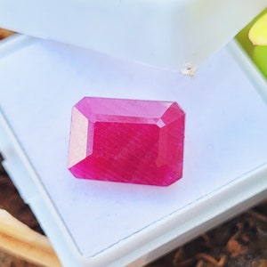 May include: A vibrant, rectangular-cut ruby gemstone with a deep pinkish-red hue and faceted surfaces. The gemstone is displayed resting inside a white, open-hinged plastic storage box, showcasing its clarity and polished edges clearly.