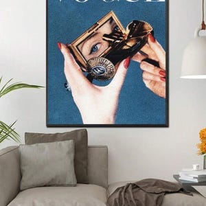 May include: A vintage Vogue magazine cover featuring a woman's hand holding a compact mirror, with the word "VOGUE" printed in white letters on a blue background.