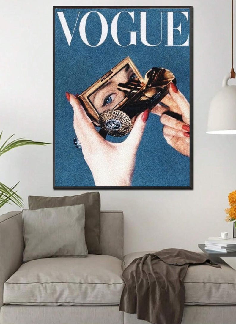 May include: A vintage Vogue magazine cover featuring a woman's hand holding a compact mirror, with the word "VOGUE" printed in white letters on a blue background.