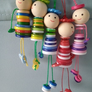 May include: Five colourful wooden dolls with button bodies and smiling faces. Each doll has a different coloured hat and is hanging from a string.