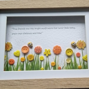 May include: A framed picture with a quote about friendship, featuring button flowers in yellow, orange, and red, with green grass. The frame is light brown.