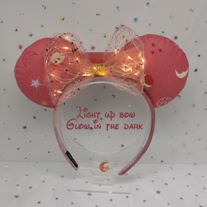Puede incluir: Diadema rosa con orejas de Minnie Mouse y un lazo iluminado. Las orejas tienen un diseño celestial con estrellas y lunas. El lazo está adornado con pequeñas luces y una superposición transparente. El texto en la diadema dice "Light up bow Glow in the Dark."