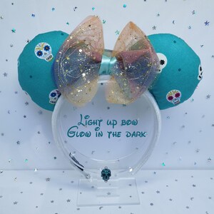 Puede incluir: Un par de orejas de ratón turquesas con diseños de calaveras de azúcar y un lazo brillante. El lazo es una mezcla de rosa, dorado y azul. El texto en el soporte de exhibición dice "Light up bow, Glow in the dark."