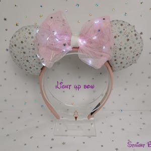 May include: A light pink headband with large ears and a bow. The ears are white with a pattern of small brown stars. The bow is light pink with small lights. The text "Light up bow" is visible.