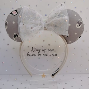 Puede incluir: Diadema de orejas de ratón grises con un lazo blanco y el texto "Light up bow, Glow in the dark". Las orejas tienen un patrón de estrellas y personajes de dibujos animados. El lazo es de tul blanco con purpurina.