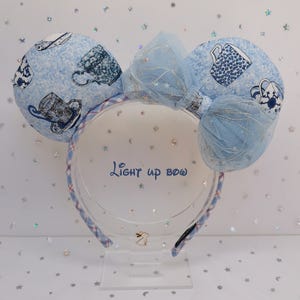 Puede incluir: Una diadema azul claro con dos orejas redondas estampadas con tazas de té. Un lazo azul a juego con detalles brillantes se encuentra entre las orejas. La diadema tiene un patrón de cuadros azules y blancos.
