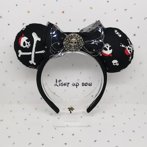 Puede incluir: Orejas de ratón negras con temática pirata con un patrón de calaveras y huesos cruzados. Las orejas tienen una diadema negra y un lazo decorativo con un emblema de calavera. El lazo está adornado con pequeñas luces. El texto "Light up bow" es visible.