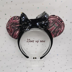 Könnte beinhalten: Schwarzes Stirnband mit zwei runden Ohren mit einem rosa-schwarzen Marmormuster. Eine schwarze Schleife mit kleinen Lichtern befindet sich oben. Der Text "Light up bow" ist auf dem Displayständer zu sehen.