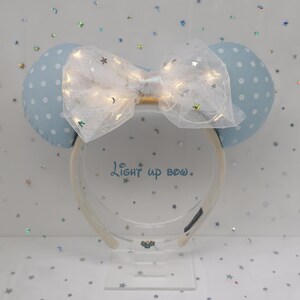 Puede incluir: Una diadema azul claro con dos orejas con lunares y un lazo blanco. El lazo está adornado con pequeñas luces parpadeantes y lentejuelas en forma de estrella. El texto "Light up bow" está impreso en el soporte transparente.