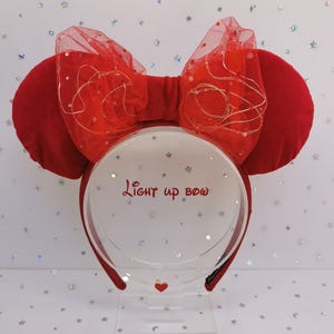 Puede incluir: Diadema roja con orejas de ratón y un lazo a juego. El lazo está hecho de red roja y tiene pequeñas luces. El texto "Light up bow" está impreso en el expositor transparente. Un pequeño corazón rojo está en la parte inferior del expositor.