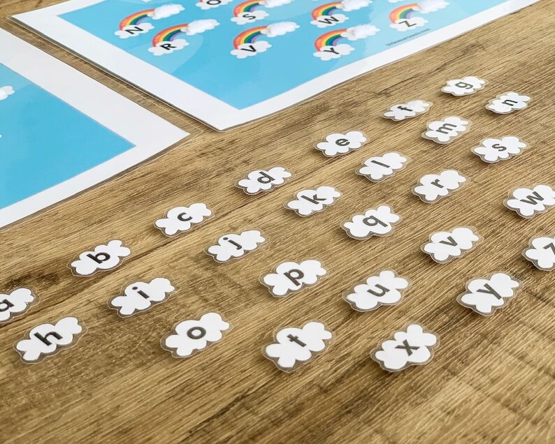 May include: A set of individual lowercase letter cutouts shaped like white clouds, each featuring a black letter. Two large rectangular sheets are visible in the background, displaying colorful rainbows above white clouds with uppercase letters.