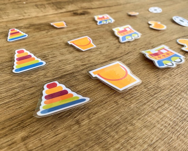May include: A set of colourful felt toy stickers, including a train, a bucket, a stack of rings, and a toy truck. The stickers are on a wooden surface.