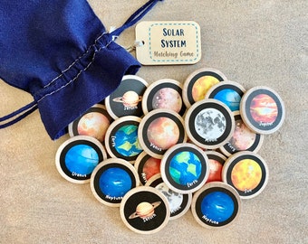 May include: A navy blue drawstring bag next to a collection of wooden discs featuring planets and celestial bodies. The discs are part of a matching game, with the words "Solar System Matching Game" on a wooden tag.