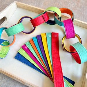 May include: A wooden tray holds a colorful felt chain and individual felt strips. The chain links are in various colors, including blue, green, orange, and pink. The felt strips are arranged in a fan shape, each with a snap closure.