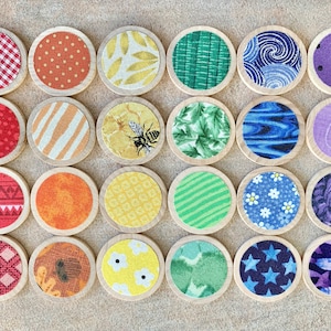 May include: A collection of twenty-four round wooden coasters with various fabric patterns. Designs include gingham, polka dots, floral, stripes, and a bee. The coasters are arranged in a grid, showcasing a variety of colors and textures.
