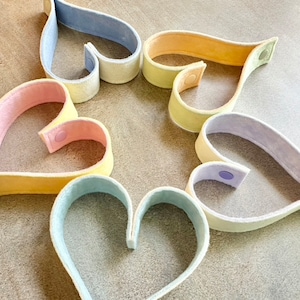 May include: A collection of heart-shaped felt bands in pastel colors, including pink, yellow, blue, and lavender. Each band has a snap closure. The arrangement suggests a craft or decorative item, possibly for a DIY project or gift.