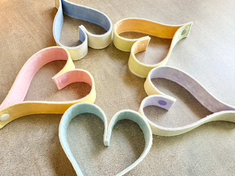 May include: A collection of heart-shaped felt bands in pastel colors, including pink, yellow, blue, and lavender. Each band has a snap closure. The arrangement suggests a craft or decorative item, possibly for a DIY project or gift.