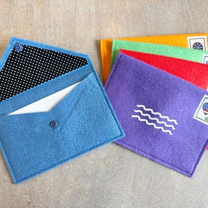 Peut inclure: Un ensemble d'enveloppes en feutrine bleu, violet, rouge, vert et orange. L'enveloppe bleue est ouverte, r&eacute;v&eacute;lant une doublure &agrave; pois noirs et blancs et une carte blanche. Chaque enveloppe ferm&eacute;e porte un cachet ondul&eacute; blanc et un timbre.