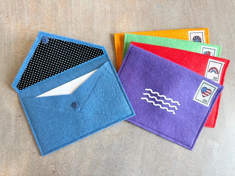 Peut inclure: Un ensemble d'enveloppes en feutrine bleu, violet, rouge, vert et orange. L'enveloppe bleue est ouverte, r&eacute;v&eacute;lant une doublure &agrave; pois noirs et blancs et une carte blanche. Chaque enveloppe ferm&eacute;e porte un cachet ondul&eacute; blanc et un timbre.