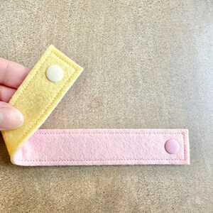 May include: Two felt straps, one yellow and one pink, connected by a white and a pink snap. The straps are stitched along the edges. The item is shown against a neutral background.