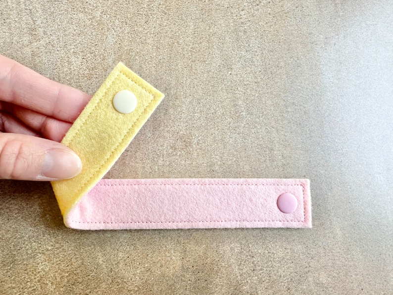 May include: Two felt straps, one yellow and one pink, connected by a white and a pink snap. The straps are stitched along the edges. The item is shown against a neutral background.