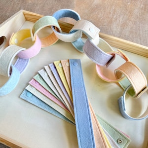 May include: Pastel-colored felt chain links and individual strips with snap closures, arranged on a light wooden tray. The chain links are in shades of pink, blue, yellow, and green. The strips are fanned out, showing the variety of colors.