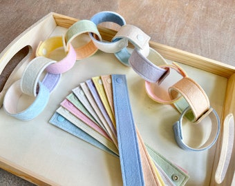 Peut inclure: Chaînes en feutre de couleurs pastel et bandes individuelles avec fermetures à bouton-pression, disposées sur un plateau en bois clair. Les chaînes sont dans des tons de rose, bleu, jaune et vert. Les bandes sont déployées, montrant la variété des couleurs.