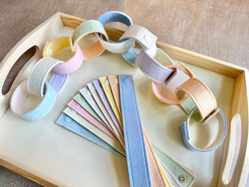 May include: Pastel-colored felt chain links and individual strips with snap closures, arranged on a light wooden tray. The chain links are in shades of pink, blue, yellow, and green. The strips are fanned out, showing the variety of colors.