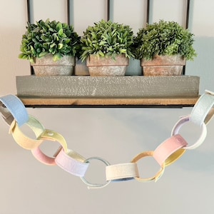 May include: A pastel-colored felt chain hangs below a shelf with three potted faux plants. The chain links are in shades of pink, yellow, blue, and white. The shelf is gray and made of wood.