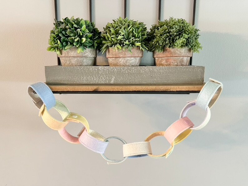 May include: A pastel-colored felt chain hangs below a shelf with three potted faux plants. The chain links are in shades of pink, yellow, blue, and white. The shelf is gray and made of wood.