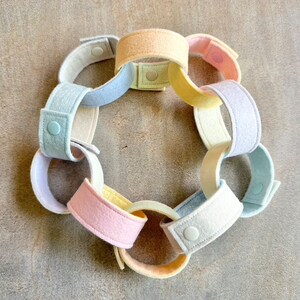 May include: A pastel-colored felt chain with interlocking loops. The chain features a variety of soft colors, including pink, yellow, blue, and cream. Each loop is connected with a snap closure. The chain is arranged in a circular shape.