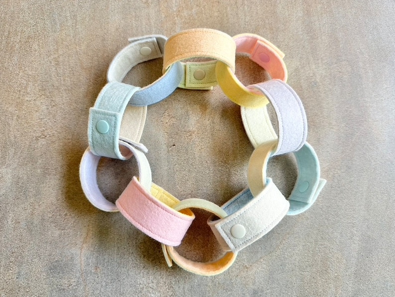 May include: A pastel-colored felt chain with interlocking loops. The chain features a variety of soft colors, including pink, yellow, blue, and cream. Each loop is connected with a snap closure. The chain is arranged in a circular shape.