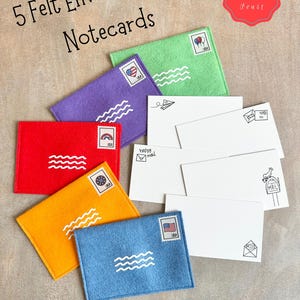 Peut inclure: Cinq enveloppes en feutrine verte, violette, rouge, orange et bleue, chacune avec un timbre blanc et des lignes ondul&eacute;es. Vingt cartes blanches pr&eacute;sentent de petites illustrations &agrave; l'encre noire. Le texte indique 5 Felt Envelopes + 20 Notecards.